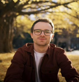 黒縁メガネをかけ、黄葉の中にいる人物写真。白いTシャツに茶色いパーカーを着ている。髪は短髪